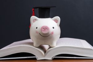 Scholarship, savings for university, cost of knowledge or financial education concept, pink smiling piggy bank with graduation hat on top of opening book with dark blackboard or chalkboard background
